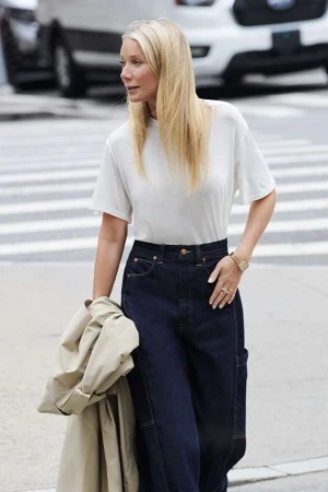 Gwyneth Paltrow wearing a white t-shirt and dark jeans holding a trench coat on a city street.