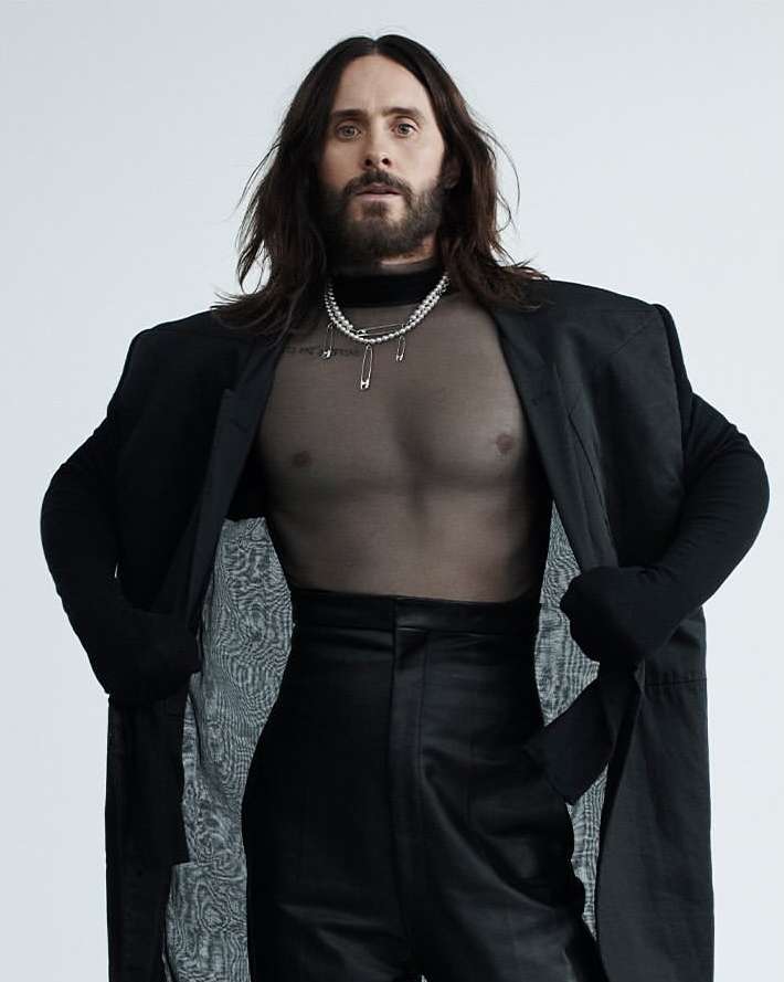 Portrait of Jared Leto wearing a black jacket and translucent top, accessorized with a pearl necklace against a white backdrop.