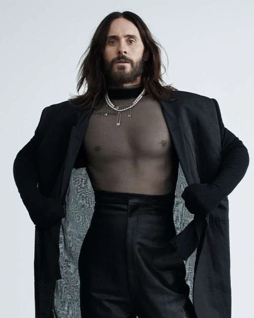 Portrait of Jared Leto wearing a black jacket and translucent top, accessorized with a pearl necklace against a white backdrop.