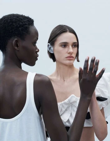 Two women in elegant white outfits pose together. One woman with dark skin stands next to a woman with fair skin in a studio setting.