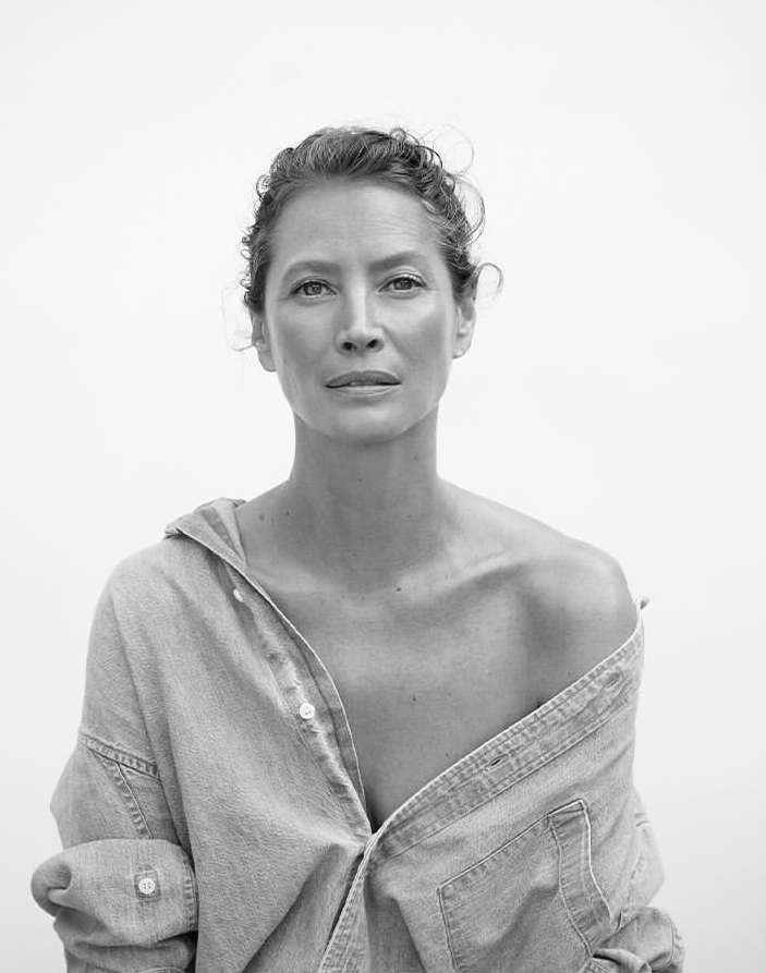 Black and white portrait of an adult female wearing an unbuttoned denim shirt, photographed at eye-level against a white backdrop.