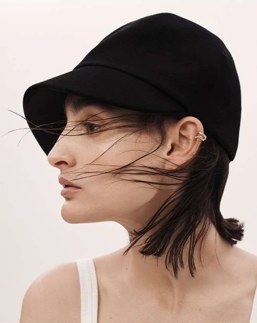 Close-up of a female model wearing a black bucket hat and a gold ear cuff on a white background in a studio fashion shot.