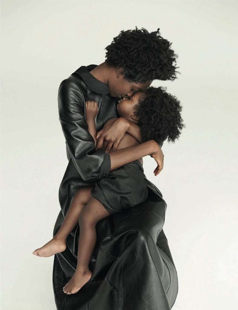 A black mother and child embrace in a tender moment, both in dark attire against a clean, bright background. A touching family portrait.