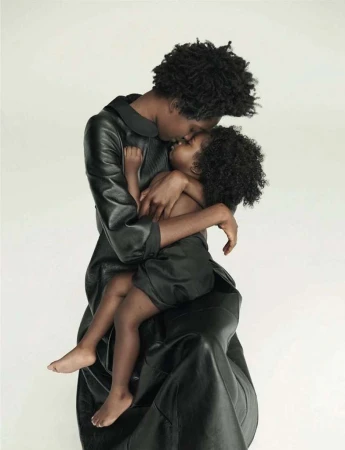 A black mother and child embrace in a tender moment, both in dark attire against a clean, bright background. A touching family portrait.