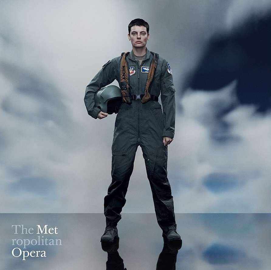 Female pilot in flight suit stands against cloudy sky, holding helmet, in full shot against a cloudy backdrop. The Met Opera text overlay.