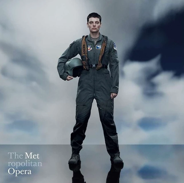 Female pilot in flight suit stands against cloudy sky, holding helmet, in full shot against a cloudy backdrop. The Met Opera text overlay.