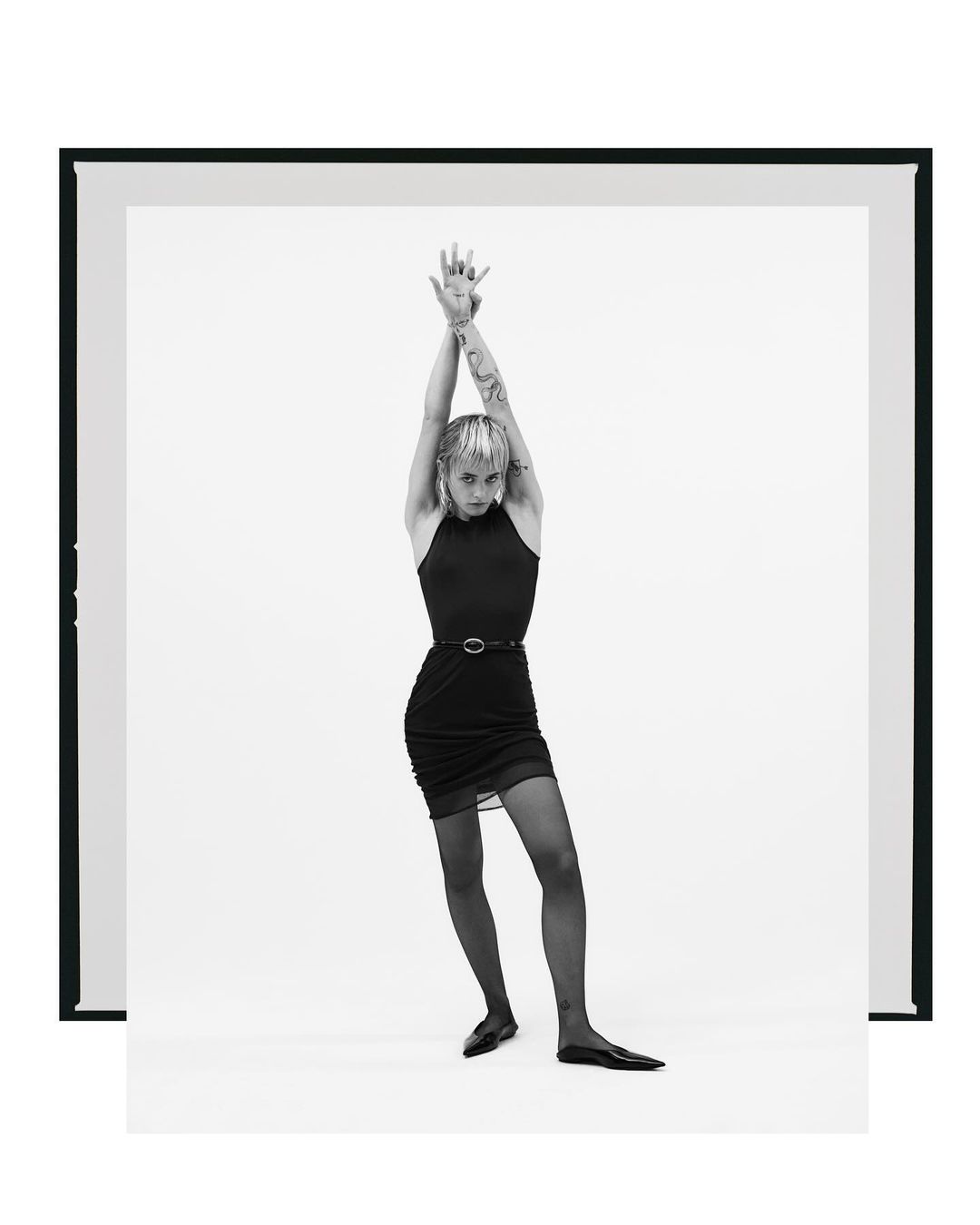 Full-length shot of a slim young woman with raised arms, wearing a black dress, tights, and ballet flats, posing in a white photo studio.