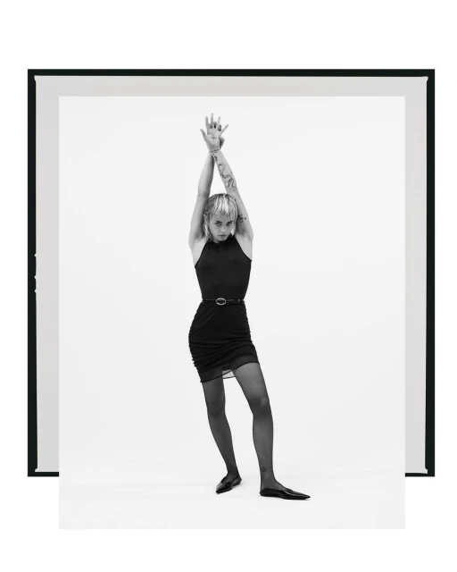 Full-length shot of a slim young woman with raised arms, wearing a black dress, tights, and ballet flats, posing in a white photo studio.