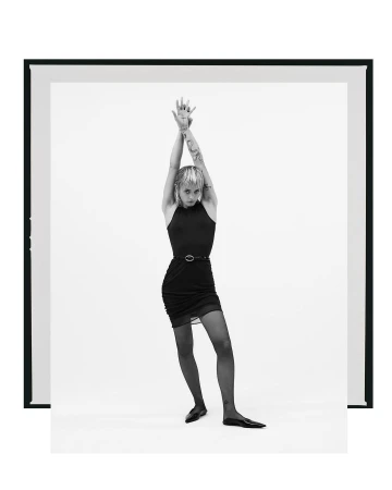 Full-length shot of a slim young woman with raised arms, wearing a black dress, tights, and ballet flats, posing in a white photo studio.