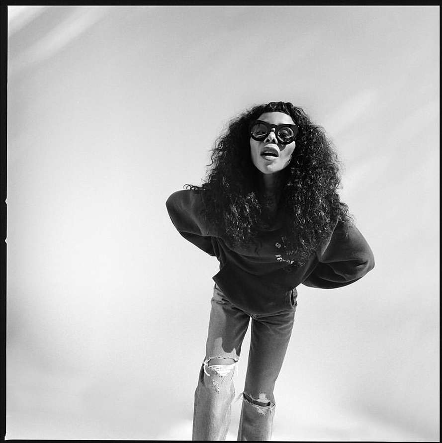 Black and white portrait of a woman with curly hair wearing sunglasses and ripped jeans by photographer Paola Kudacki