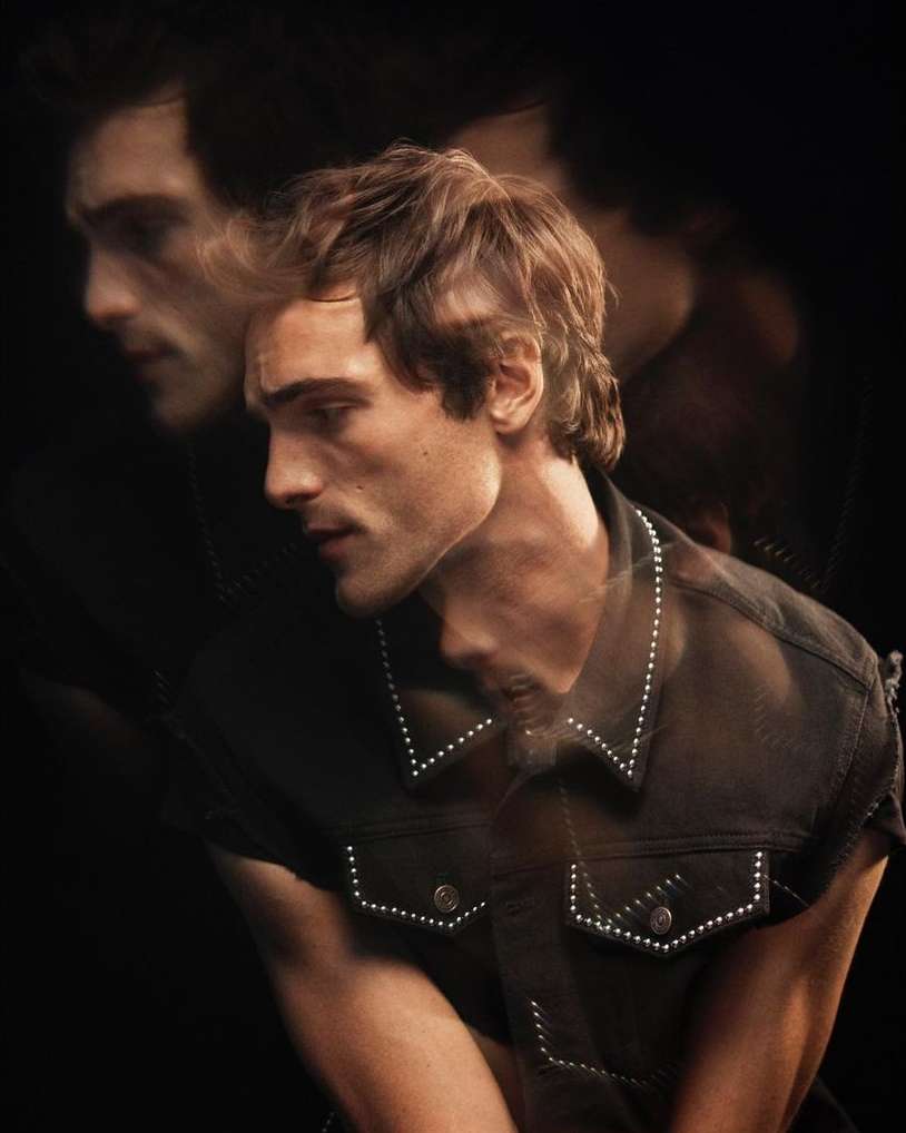 Portrait of a young adult male in a sleeveless denim jacket, set against a dark background with a blurred effect, studio lighting.