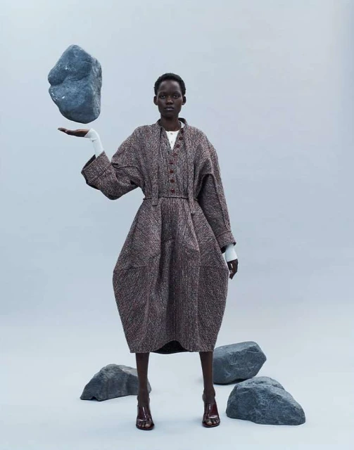 Fashion shot of a woman in a sculptural coat holding a rock in her outstretched hand, set against a light blue background in a studio.