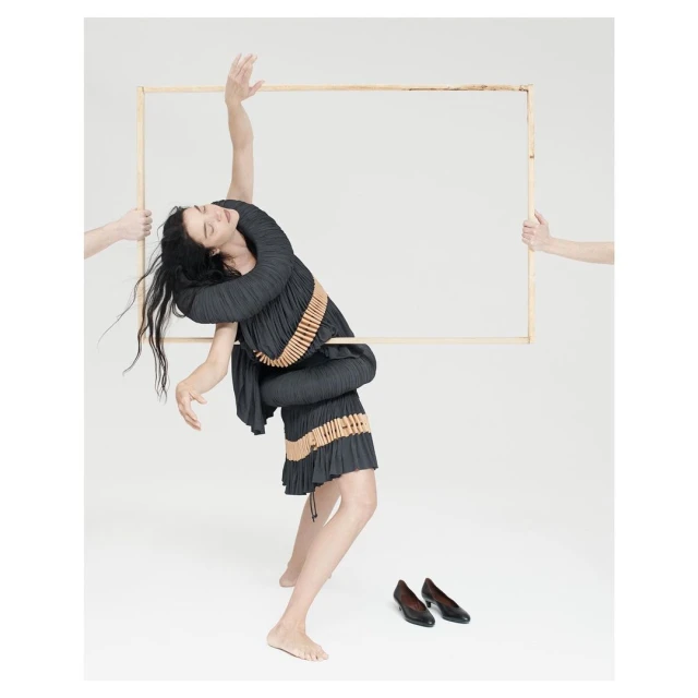 Fashionable woman in pleated dress poses with a wooden frame held by disembodied arms, with black shoes on a white seamless backdrop.