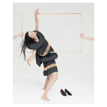 Fashionable woman in pleated dress poses with a wooden frame held by disembodied arms, with black shoes on a white seamless backdrop.