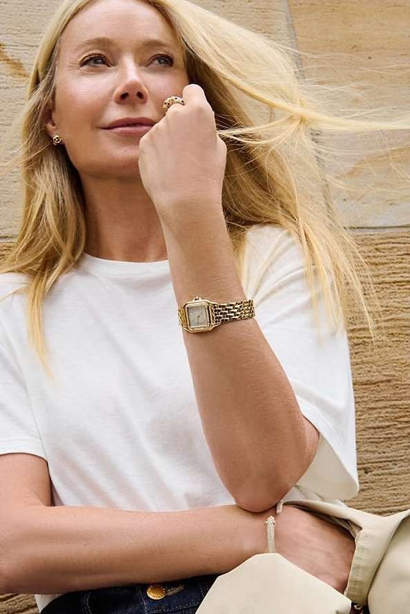 Gwyneth Paltrow wearing a white t-shirt and gold watch outdoors against a textured stone wall