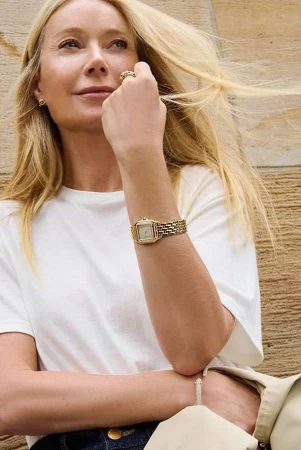 Gwyneth Paltrow wearing a white t-shirt and gold watch outdoors against a textured stone wall