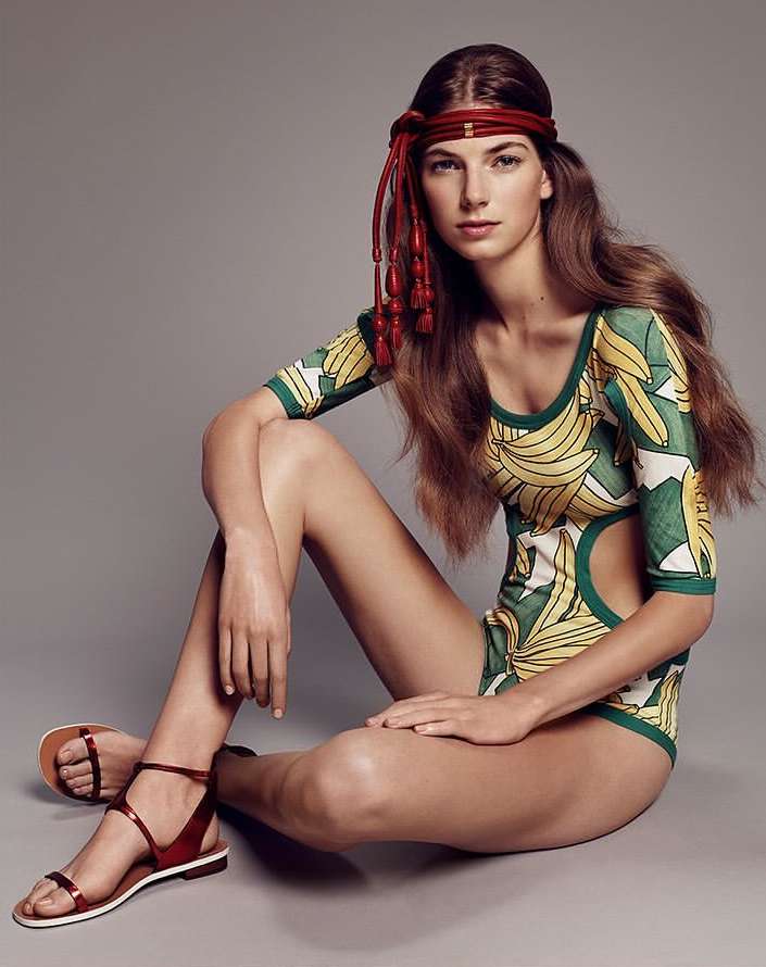 Woman in a green banana-print bodysuit and red sandals sits in a studio with a grey backdrop, showcasing a fashion-forward summer style.