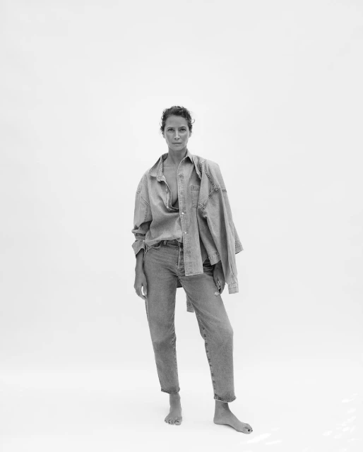 Monochrome full shot of an adult woman standing barefoot in light denim jeans and an oversized denim shirt against a white background.