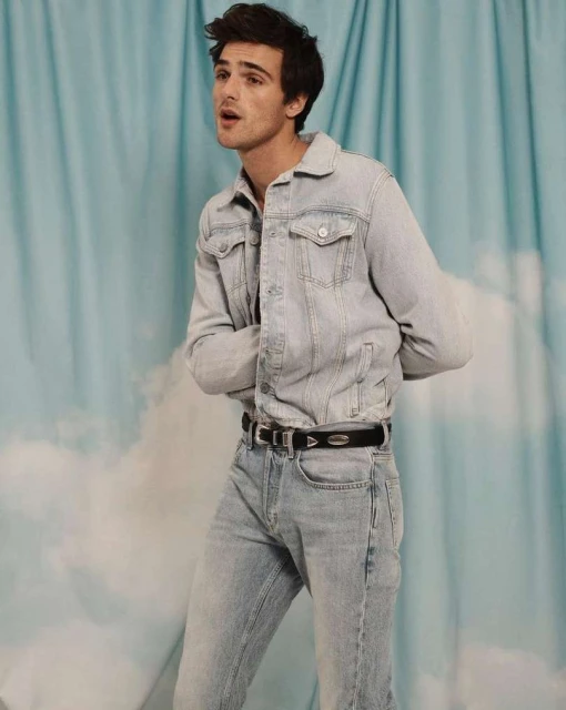 Adult male in denim jacket and jeans poses against a light blue backdrop with cloud accents in a medium shot, looking off camera.