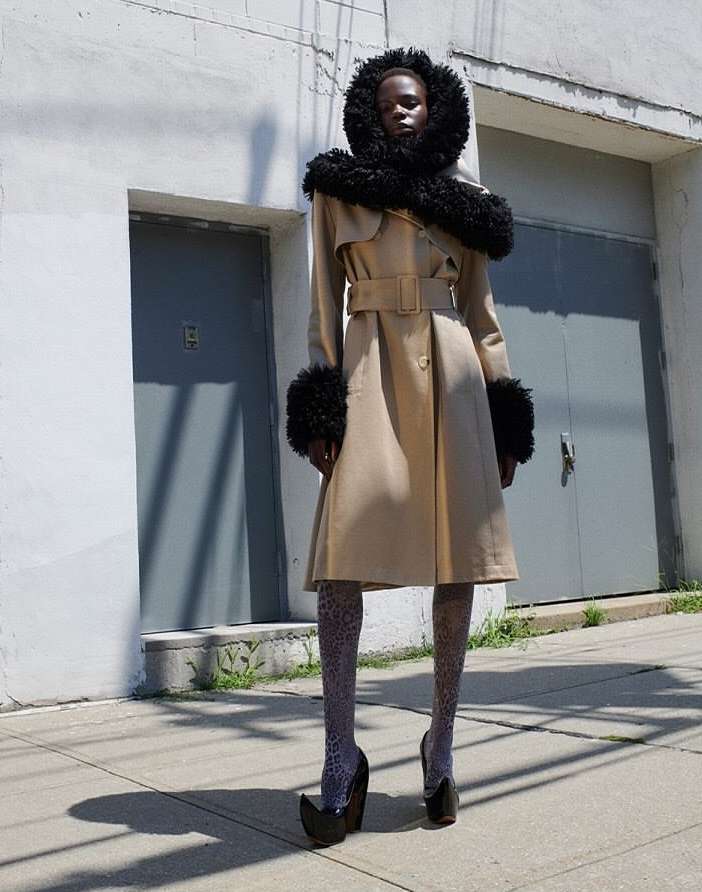 Fashion model wears a trench coat with fur trim, patterned tights, and platform heels standing outdoors on a sunny day in an urban setting.