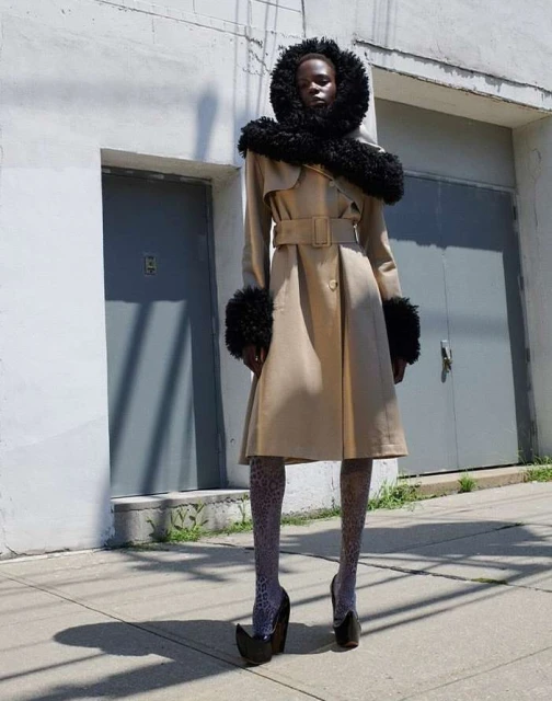 Fashion model wears a trench coat with fur trim, patterned tights, and platform heels standing outdoors on a sunny day in an urban setting.