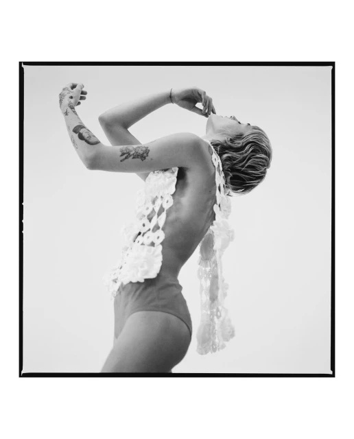 Black and white studio portrait of a young adult woman posing with arms up, wearing a crochet top and swimsuit bottoms on a white backdrop.