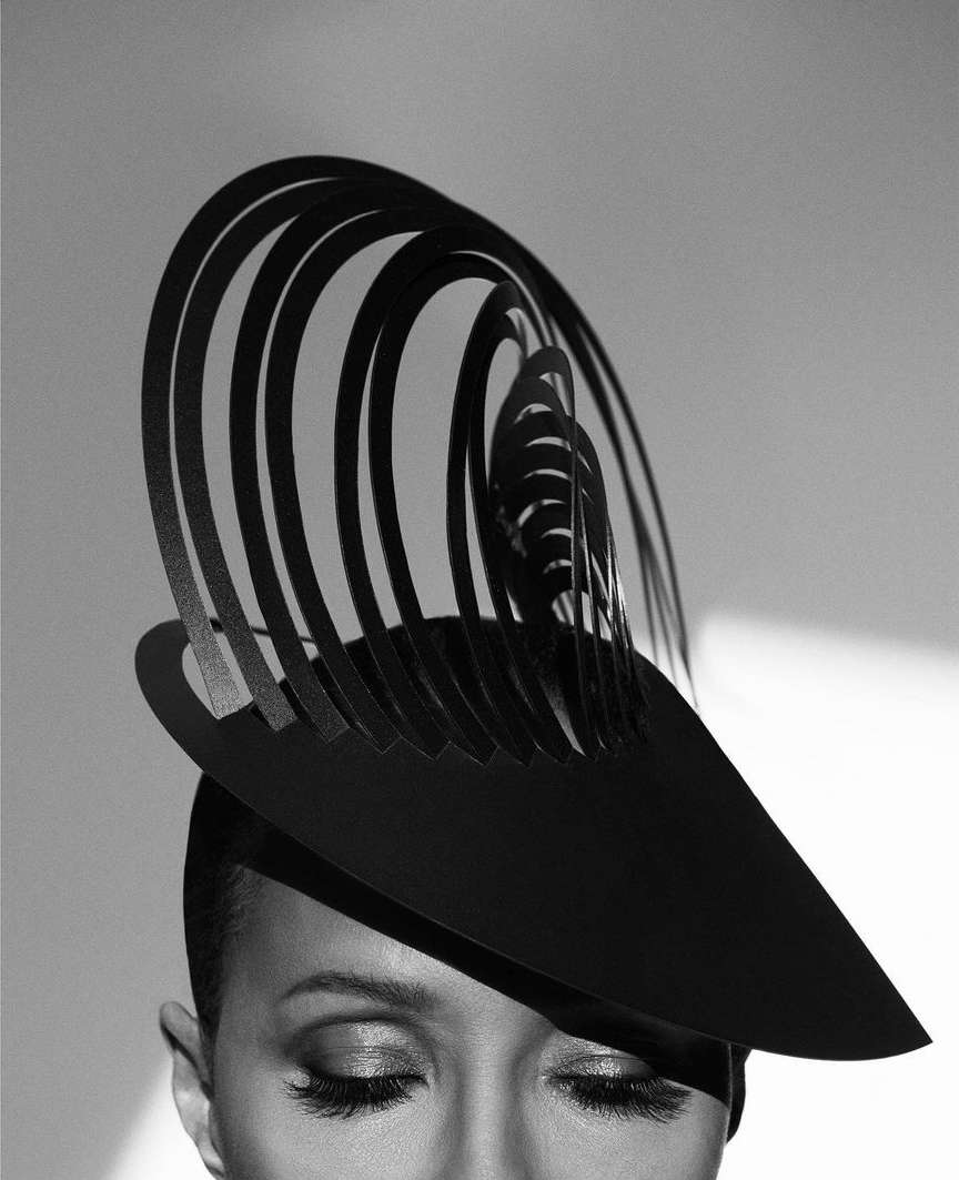Black and white portrait of a woman with a modern sculptural hat featuring geometric, curved designs, styled with dramatic eye makeup.