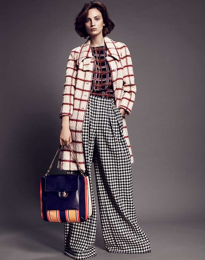 Fashion model poses in a studio with a plaid coat, shirt, and pants, while holding a handbag against a grey backdrop.