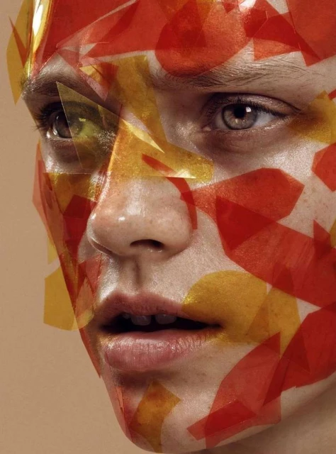 Close-up portrait of a person's face covered in translucent red and yellow geometric shapes, set against a neutral background in studio lighting.