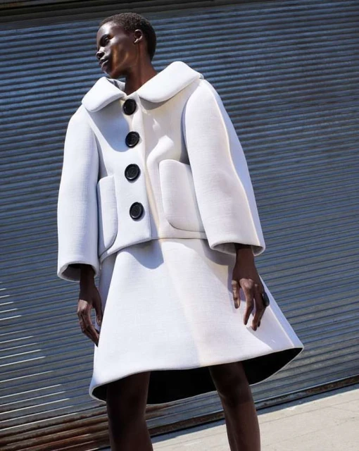 Fashion shot of an adult woman in a white skirt suit posing outdoors against a gray metal wall in sunlight for editorial fashion.