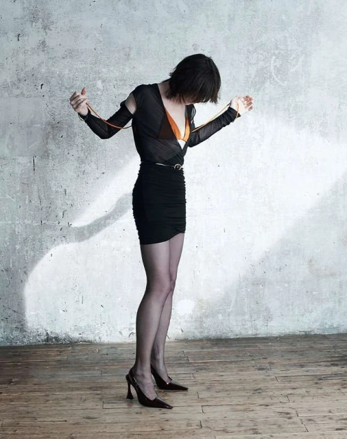 A woman with short dark hair poses in a black mini dress and heels in front of a distressed grey wall, looking down with her arms slightly raised.