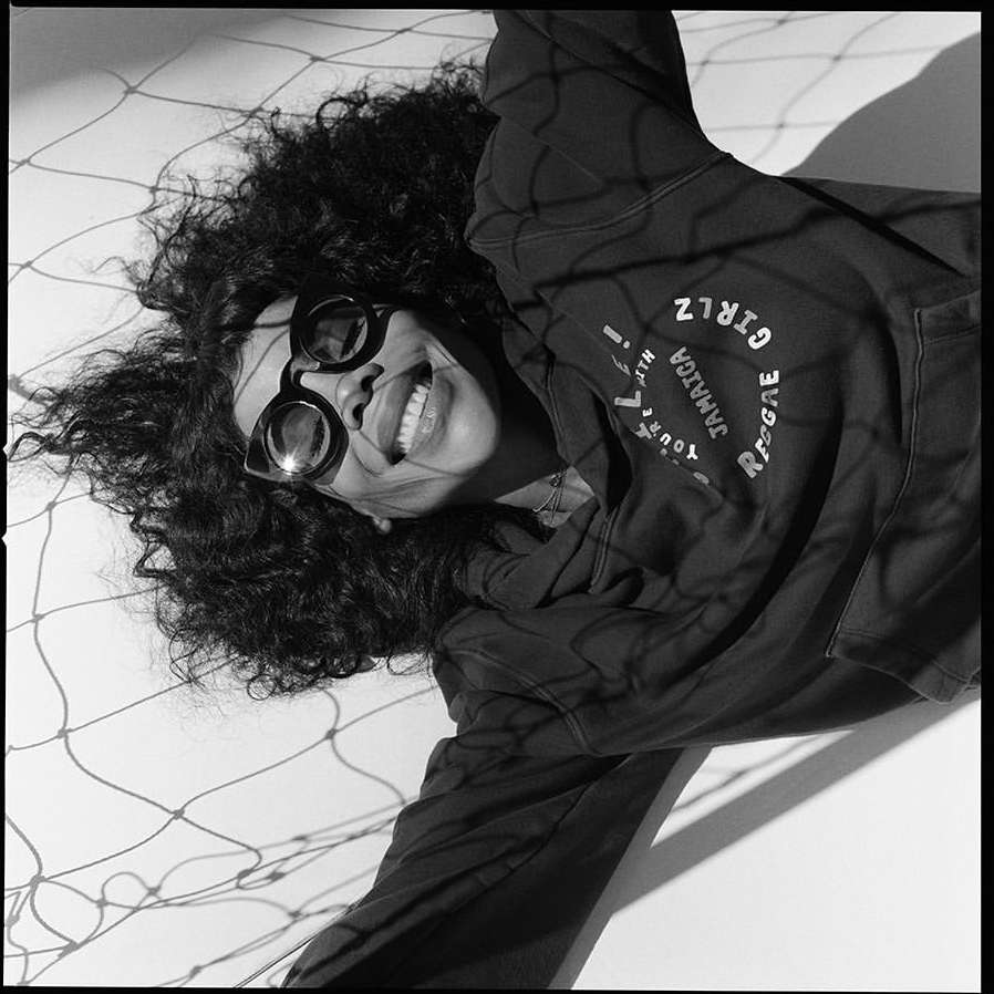 Black and white portrait of a woman with curly hair wearing round sunglasses and a hoodie, lying on a white surface with net shadows.