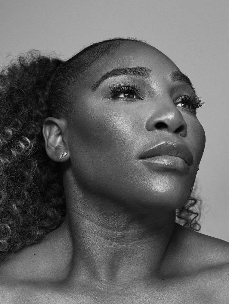 Striking black-and-white portrait of Serena Williams, face forward, eyes gazing upwards, stylish curly hair pulled back into a ponytail.