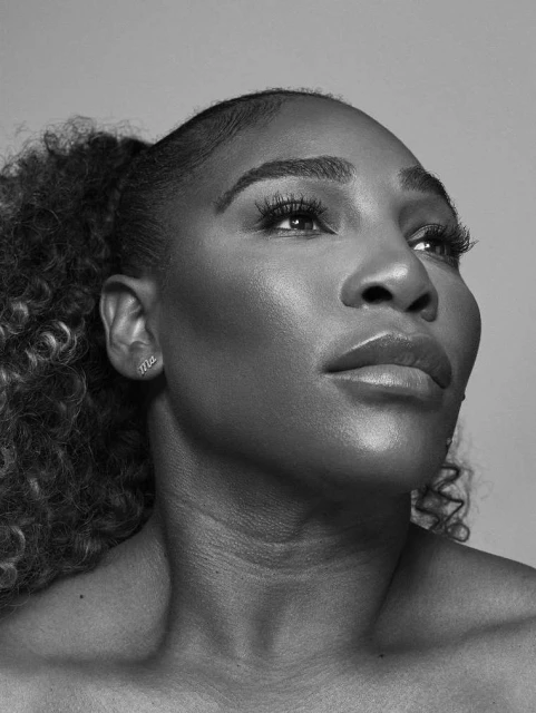 Striking black-and-white portrait of Serena Williams, face forward, eyes gazing upwards, stylish curly hair pulled back into a ponytail.