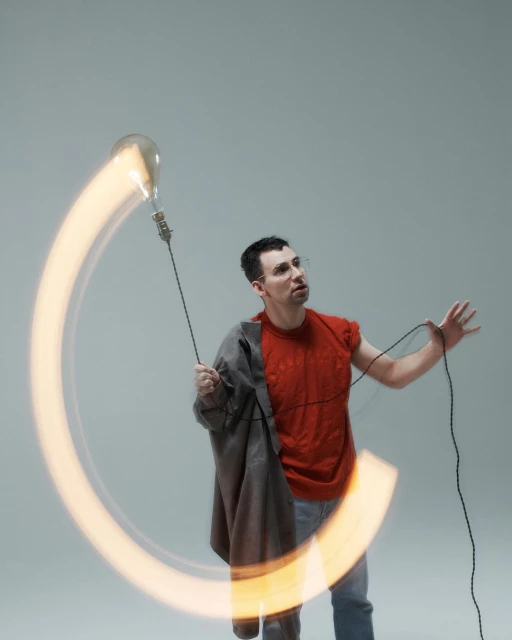 Adult male poses in photo studio with lightbulb creating a light trail effect. He is holding power cord of light in indoor environment.