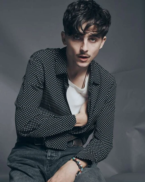 Close-up studio portrait of a young adult male with dark hair and a mustache, wearing an open patterned shirt, jeans, and a beaded bracelet.