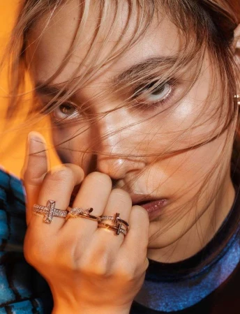 Close-up portrait of a woman wearing diamond rings on her fingers, with blonde hair falling across her face against an orange background.