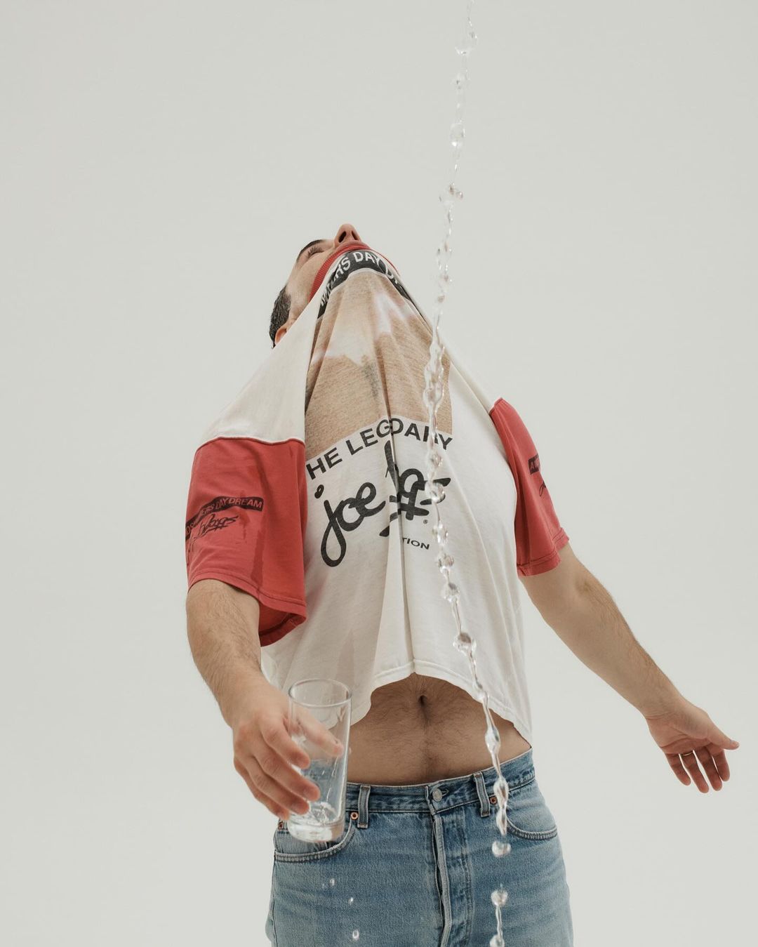Adult male in jeans and a t-shirt has water poured on his head from above, holding an empty glass, against a white backdrop.