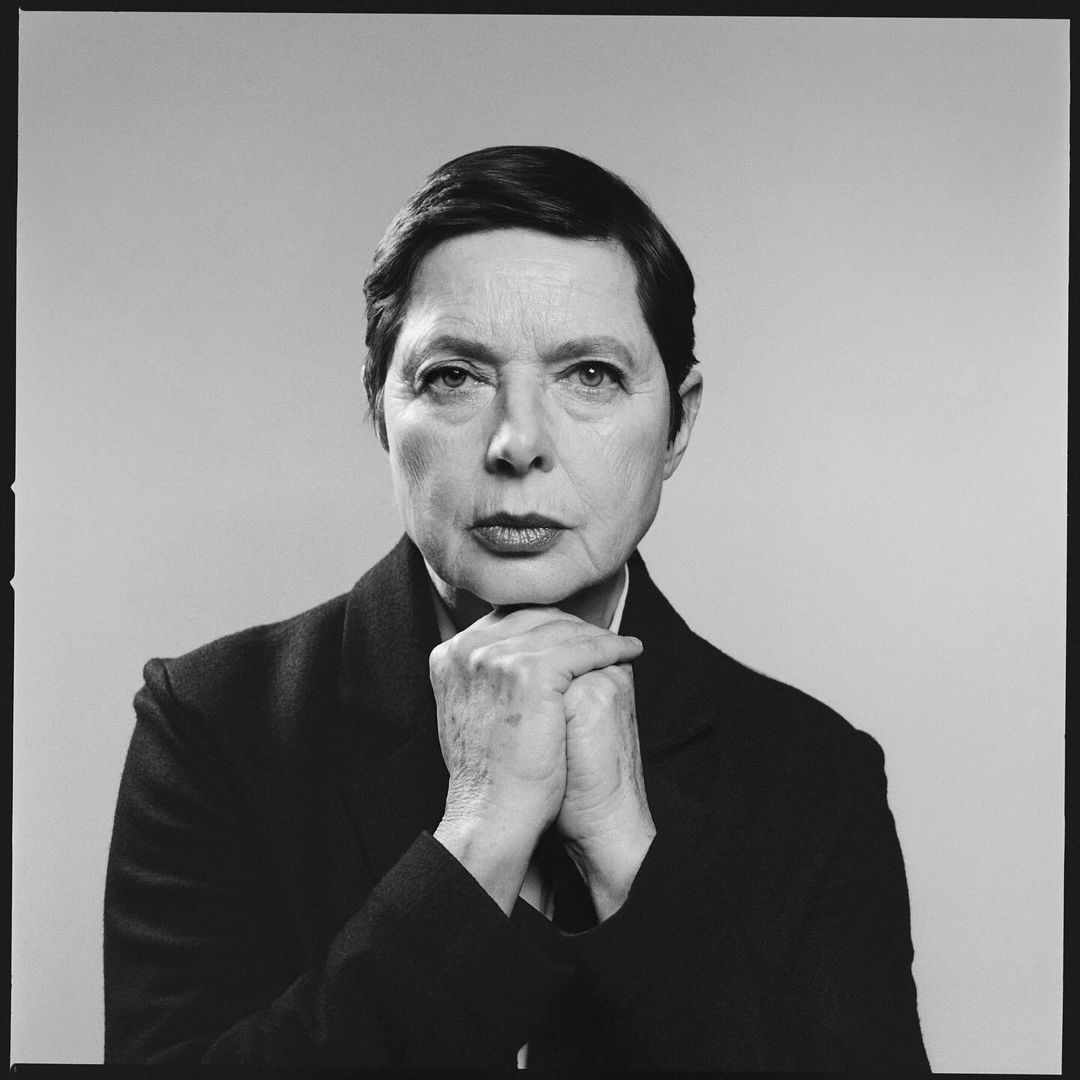 Portrait of Isabella Rossellini with hands clasped beneath chin wearing a dark blazer against a grey backdrop by Paola Kudacki.