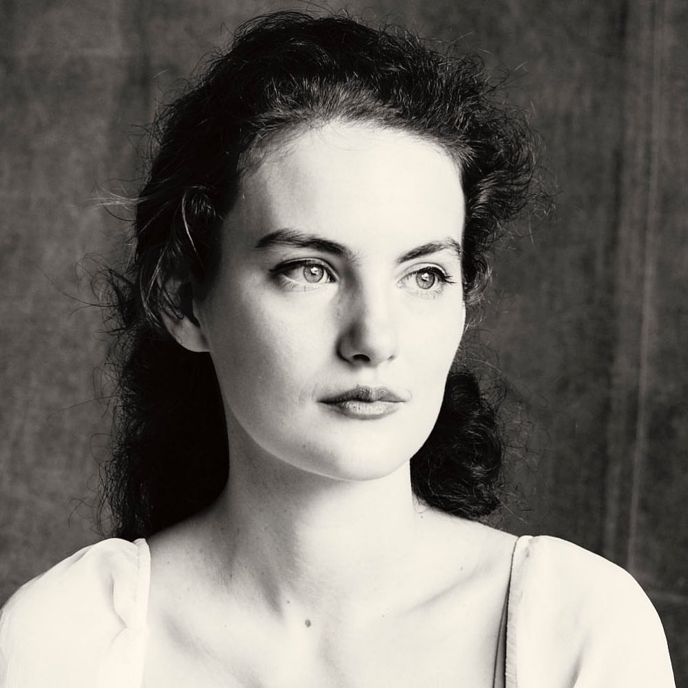 Monochrome portrait of a young adult woman with dark curly hair looking to the side against a dark blurred background in a studio setting.