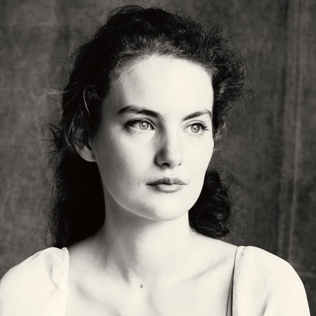 Monochrome portrait of a young adult woman with dark curly hair looking to the side against a dark blurred background in a studio setting.