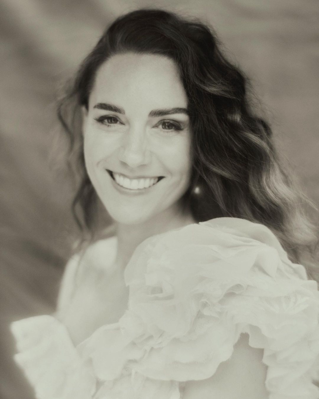 Close-up portrait of a smiling adult woman with wavy dark hair wearing a white ruffled top against a blurred neutral background.