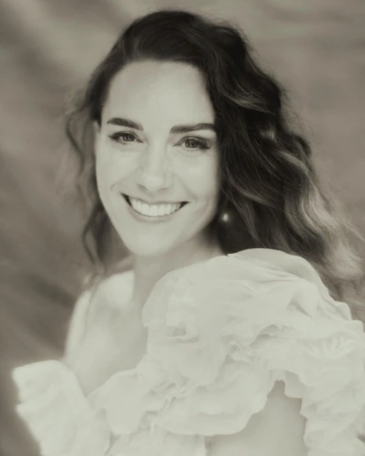 Close-up portrait of a smiling adult woman with wavy dark hair wearing a white ruffled top against a blurred neutral background.