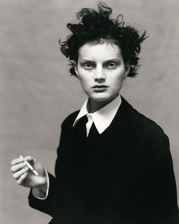 Monochrome portrait of a person with short curly hair wearing a dark suit jacket and white collared shirt holding a cigarette.
