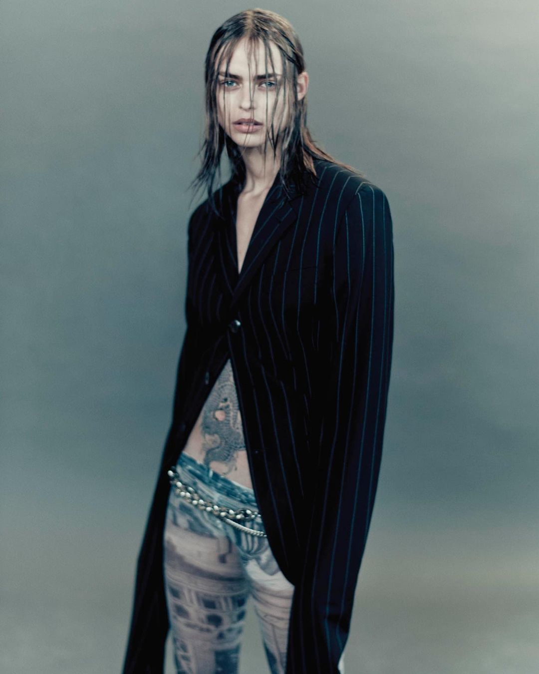 A slender woman with wet hair in a pinstripe jacket and patterned pants poses in a studio with a grey backdrop.