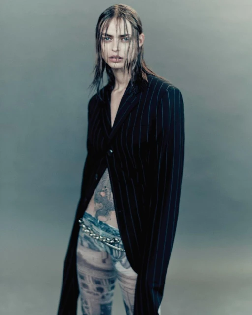 A slender woman with wet hair in a pinstripe jacket and patterned pants poses in a studio with a grey backdrop.