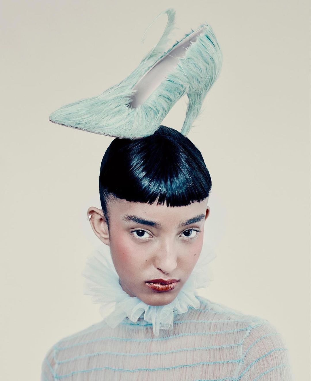 Fashion portrait of a young adult with dark hair wearing a sheer aqua top and a shoe as a hat, set against a neutral background.