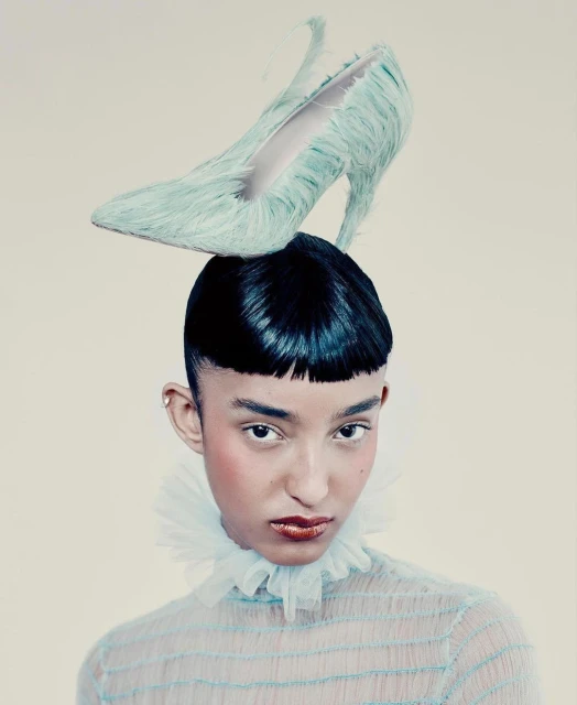 Fashion portrait of a young adult with dark hair wearing a sheer aqua top and a shoe as a hat, set against a neutral background.