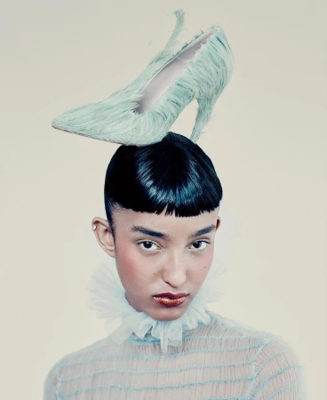 Fashion portrait of a young adult with dark hair wearing a sheer aqua top and a shoe as a hat, set against a neutral background.