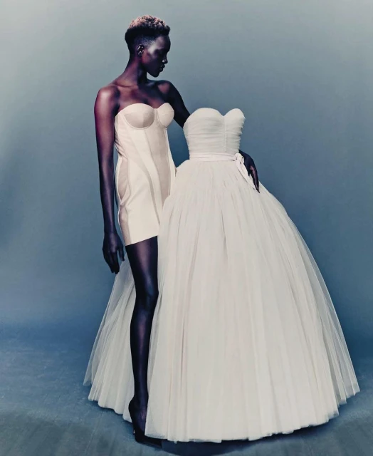 Fashion editorial shot of a slim African woman in a white bustier dress, standing beside a large tulle ball gown against a blue background.